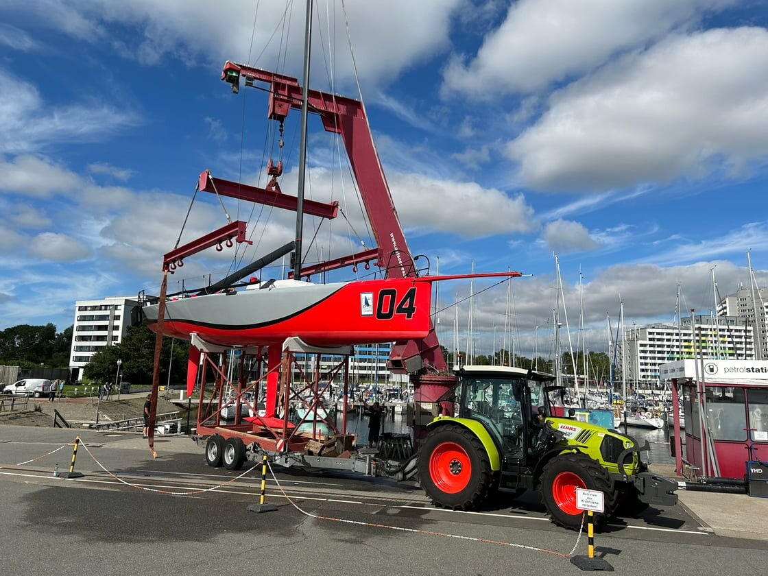 Roter Hafenkran hebt eine weiße Segelyacht, im Vordergrund grüner Traktor, blauer Himmel mit Wolken.